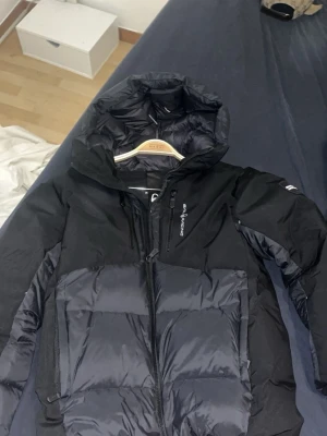 Svart pufferjacka från  sail racing - Snygg svart pufferjacka från sail racing med huva och dragkedja framtill. Jackan har en stilren design med diskret logga på bröstet och stora, vadderade paneler som håller dig varm. Perfekt för kalla dagar och enkel att matcha med olika outfits.