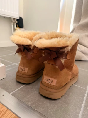  UGG moon boots med rosett - Säljer ett par klassiska bruna UGG moon boots i mocka med fluffigt beige foder och söta bruna rosetter bak. Skorna har en robust sula och är perfekta för kalla dagar. UGG-logga bak på hälen och mjuk insida för extra komfort. Storlek 38