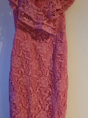 Rosa spetsklänning med volang - Säljer en rosa spetsklänning med vackert blommönster och volangdetalj upptill. Klänningen har en figurnära passform och är knälång, perfekt för dig som vill ha en romantisk och feminin look. Materialet är spets som ger en exklusiv känsla.