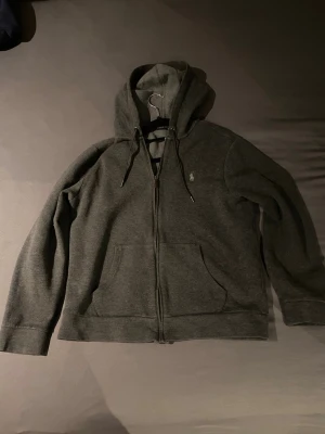 Mörkgrå hoodie från Polo Ralph Lauren - Mörkgrå hoodie från Polo Ralph Lauren med dragkedja framtill, två stora fickor och justerbar huva med snören. Liten broderad logga på bröstet. Tillverkad i mjukt bomullsmaterial som känns skönt mot huden. Perfekt för en avslappnad och stilren look.