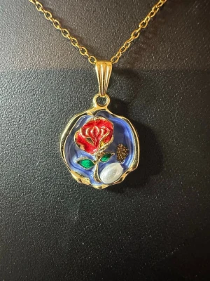 Halsband med ros och pärldetalj - Unikt halsband med guldfärgad kedja och runt hänge. Hänget har en röd ros med gröna blad, blå bakgrund och en vit pärldetalj. Detaljer i guld och små inslag av brunt ger en konstnärlig känsla. Perfekt för dig som gillar färgstarka accessoarer.
