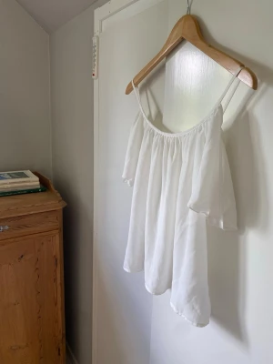 Vit off shoulder topp med tunna band - Superfin vit offshoulder blus med tunna axelband och vida, luftiga ärmar. Tyget är lätt och har en snygg fallande effekt. Perfekt för en fräsch och stilren look. Passar dig som gillar en luftig och avslappnad vibe. Var tvungen att klippa av lappen då den kliade. Storlek S/M