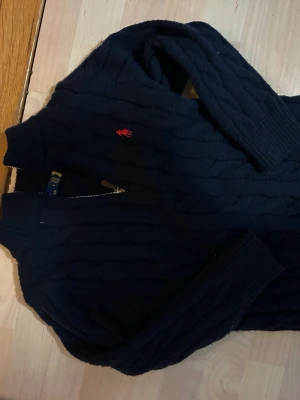 Mörkblå kabelstickad tröja Polo Ralph Lauren pris kan diskuteras!!! - Mörkblå kabelstickad tröja från Polo Ralph Lauren med dragkedja och röd broderad logga på bröstet. Tröjan har hög krage, långa ärmar och ribbade muddar. Perfekt för dig som gillar klassisk och stilren look.