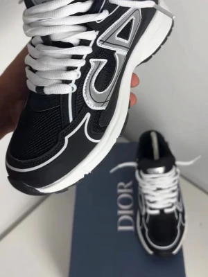 Svarta Dior sneakers med vita detaljer - Snygga svarta Dior sneakers med vita och silverfärgade detaljer samt tjock vit sula. Skorna har tydliga CD-loggor på sidan och vita skosnören. Ovandelen är i mesh och läder för en modern och sportig look.