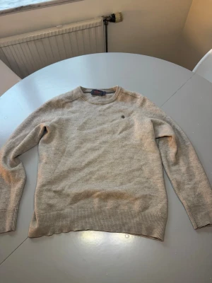 Beige merinoull tröja från Morris - Snygg beige stickad tröja från Morris med rund halsringning och diskret broderad logga på bröstet. Tröjan har ribbade muddar vid ärmslut och nederkant, perfekt för en stilren och enkel look.