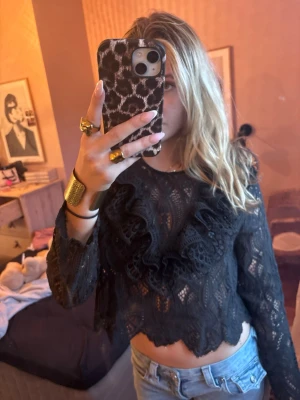 Svart spetsblus med volanger - Trendig svart spetsblus med långa ärmar och snygga volangdetaljer framtill. Blusen har genombrutet spetsmönster och rund halsringning. Perfekt att styla med jeans eller kjol för en cool look.