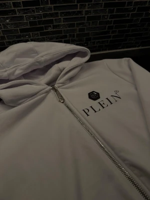 Philipp Plein hoodie  - Snygg vit hoodie från Philipp Plein med dragkedja framtill och svart logga på bröstet. Hoodien har en klassisk huva och är tillverkad i ett mjukt material. Perfekt för dig som vill gå ut med klass 
