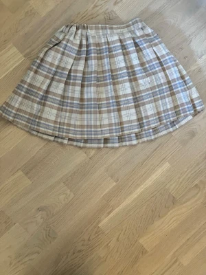 Rutig kjol - Snygg kortkjol i beige, brunt och blått med rutigt mönster och veckad design. Kjolen har bred linning och hällor för bälte, perfekt för en trendig och avslappnad stil. Passar till både sneakers och boots.