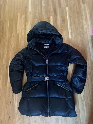 Svart lång dunjacka med huva - Snygg svart dunjacka med huva och bälte i midjan. Jackan har två stora snedställda fickor med dragkedja och en markerad midja för en schysst siluett. Perfekt för kalla vinterdagar och riktigt varm tack vare det tjocka dunfodret.