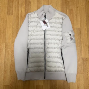Ljusgrå/beige cardigan från Moncler - Snygg ljusgrå/beige cardigan från Moncler med quiltad front i blankt material och stickade ärmar. Tröjan har hög krage, två svarta dragkedjor framtill och en ficka med Moncler-logga på ärmen. Perfekt för dig som gillar exklusiv streetwear.
