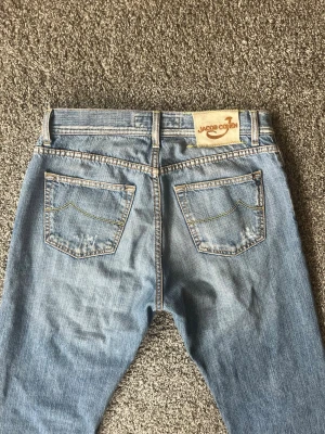 Ljusblå jeans från Jacob Cohen - Säljer ett par ljusblå jeans från Jacob Cohen med klassisk femficksmodell och raka ben. Jeansen har snygga kontrastsömmar och en patch med logga bak i midjan. Perfekta för en avslappnad och trendig stil.