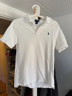 Vit pikétröja från Ralph Lauren - Klassisk vit pikétröja från Ralph Lauren med korta ärmar och krage. Tröjan har två knappar vid halsen och den ikoniska broderade loggan på bröstet. Tillverkad i mjuk bomull som känns skön mot huden. Perfekt till jeans eller shorts.