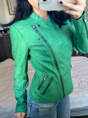 Grön skinnjacka med dragkedjor - Snygg grön skinnjacka med flera dragkedjor framtill och vid fickorna. Jackan har en figurnära passform och ståkrage med knappdetaljer. Perfekt för dig som vill sticka ut med en färgstark look.