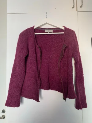 Vinröd kofta med dragkedja - Mysig lila kofta med lång ärm och dragkedja framtill. Koftan har en rund halsringning och är gjord i ett fluffigt, stickat material som känns både trendigt och bekvämt. Perfekt att slänga på sig när det är lite kyligare ute.