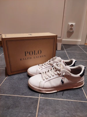 Vita Polo Ralph Lauren sneakers - Snygga vita sneakers från Polo Ralph Lauren med klassisk låg profil. Skorna har vita snören, diskret logga på sidan och detaljer i marinblått och rött på hälen och snörningen. Tillverkade i slätt läder för en clean och stilren look.