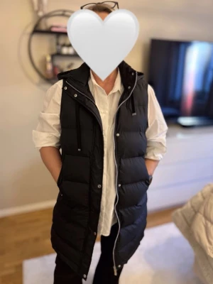 Svart lång dunväst med huva - Säljer min svarta väst då den inte kommer till användning! Super fin och skön nästan aldrig använd så den är i super skick💕