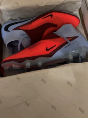 Nike Phantom Vision fotbollsskor - Säljer ett par Nike Phantom Vision fotbollsskor i en snygg röd och ljusblå färgkombination. Skorna har en strumpliknande passform utan snörning, med svarta detaljer och robusta dobbar för bra grepp på planen. Perfekta för dig som vill ha kontroll och stil på fotbollsplanen.