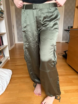 Olivgröna satincargobyxor - Säljer ett par olivgröna cargobyxor i glansigt satinmaterial. Byxorna har breda ben, stora fickor på sidorna och dragsko vid benslut för en cool look. Perfekta för dig som vill ha en trendig och avslappnad stil. Ny med prislapp. Jättefint skick. Aldrig använda.