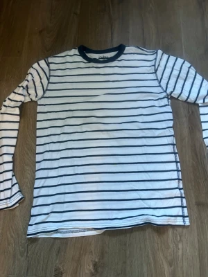 Randig långärmad topp från Cubus - Vit långärmad topp från Cubus med tunna marinblå horisontella ränder och mörkblå kant vid halsen. Tillverkad i mjuk bomull, perfekt för en avslappnad och stilren look. Klassisk passform och enkel att matcha med olika outfits.