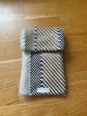 Beige och brun vävd plånbok - Unik handvävd plånbok från Botkyrka i beige, brunt och inslag av lila. Plånboken har ett vikbart lock och ett rutigt samt diagonalt mönster. Tillverkad i textilmaterial med en tydlig etikett på framsidan. Perfekt för dig som gillar hantverk och stilrena detaljer.