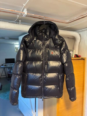 Mörkblå dunjacka från Polo Ralph Lauren - Säljer en mörkblå, glansig dunjacka från Polo Ralph Lauren med huva och dragkedja. Jackan har flera fickor med dragkedja och den klassiska röda loggan på bröstet. Perfekt för kalla vinterdagar och riktigt snygg streetstil. Köpte den i november förra året så är bara använd i knappt 4 månader!