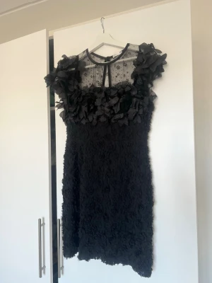 Svart blommig kortklänning Ida Sjöstedt - Supersnygg svart kortklänning från Ida Sjöstedt med 3D-blommor och transparent mesh upptill. Klänningen har rund hals, voluminösa axlar och blommiga detaljer över hela plagget. Perfekt för dig som vill sticka ut med en unik look.