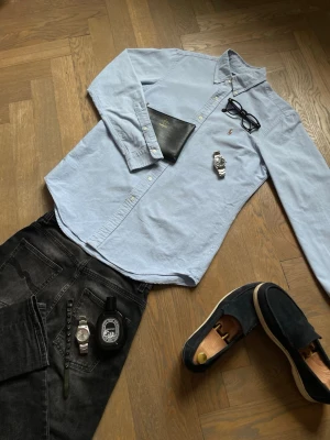 Ljusblå skjorta från Ralph Lauren - Klassisk ljusblå skjorta från Ralph Lauren med button-down krage och broderad logga på bröstet. Skjortan har långa ärmar, normal passform och är tillverkad i mjuk bomull. Perfekt för en clean och stilren look. Storlek M