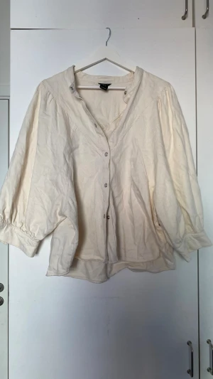 Krämvit blus med tryckknappar - Luftig krämvit blus från Lindex med trekvartsärmar och snygga tryckknappar framtill. Blusen har en avslappnad passform och är tillverkad i ett mjukt, vävt bomullstyg. Perfekt för en trendig och bekväm look.