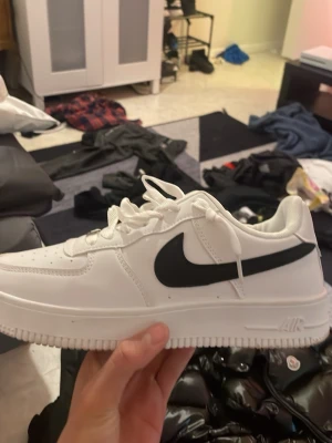 Nike Air Force 1 vita sneakers - Klassiska vita Nike Air Force 1 sneakers med svart swoosh på sidan. Skorna har en låg silhuett, vita skosnören och en robust sula med Air-dämpning. Tillverkade i läder med detaljer i gummi och textil. Perfekta för dig som gillar stilrena och tidlösa sneakers.