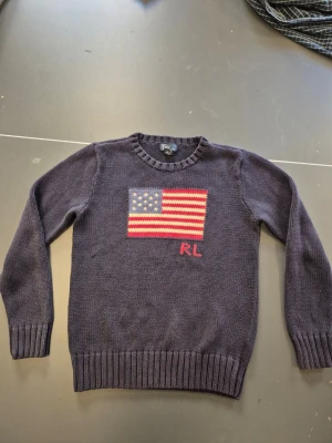 Mörkblå stickad tröja Polo Ralph Lauren - Mörkblå stickad tröja från Polo Ralph Lauren med amerikansk flagga och RL-broderi på bröstet. Tröjan har rund halsringning och ribbade muddar vid ärmslut och nederkant. Perfekt för dig som gillar klassisk. GAMLA modellen som gör den värd mer limited edition