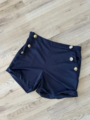 Marinblå shorts med guldknappar storlek S - Snygga marinblå shorts med sex dekorativa guldknappar framtill. Shortsen har hög midja och är sydda i ett mjukt, lätt material som ger en stilren look. Perfekta för sommardagar när du vill ha något både bekvämt och trendigt.