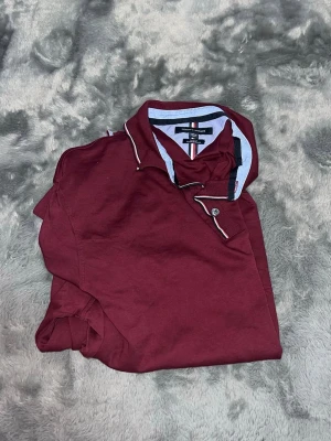 Tommy Hilfiger Slim Fit Polo – Burgundy - en riktigt fräsch pikétröja från Tommy Hilfiger i en snygg vinröd färg. Den har snygga kontrastränder vid kragen och knapparna som ger det där lilla extra. • Passform: Slim Fit. Sitter riktigt snyggt och följsamt på kroppen. • Detaljer: Klassisk broderad flagga på bröstet och detaljerad krage. • Skick: Toppskick, precis nytvättad och redo för användning! 