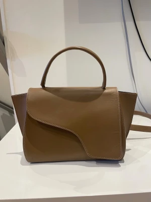 Brun handväska i läder med unik form - Säljer denna handväskan från ATP. Inte mycket använd och därav i bra skick. Inga skråmor än det som syns på bilderna. Den är i den större modellen. Nypris: 5200kr Hör av er vid funderingar💗