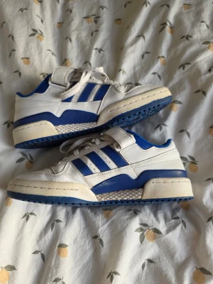 Adidas Forum Low sneakers - 41 ⅓ - Säljer ett par Adidas Forum Low sneakers i vitt och blått läder. Skorna har kardborreband upptill, blå yttersula och retroinspirerad design. Perfekta för dig som gillar streetwear och sportig stil.