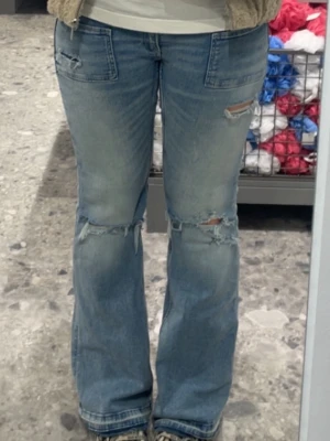 Ljusblå slitna bootcut jeans - Säljer ett par ljusblå bootcut jeans med slitningar och hål vid knäna och låren. Jeansen har framfickor och en avslappnad passform. Perfekta för en trendig och avslappnad stil. Jag har sprättat upp byxbenen. 