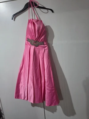 Rosa halterneck klänning med pärldetalj - Snygg rosa halterneck klänning med veckad kjol och markerad midja med pärlbroderad detalj framtill. Klänningen har en glansig finish och knytband i nacken, perfekt för speciella tillfällen.