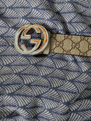 Beige G G-bälte från  - Säljer ett klassiskt beige bälte från G G med det ikoniska G G-mönstret och en stor, rund metallspänne i silver och guld. Perfekt accessoar för att lyfta vilken outfit som helst. Bältet har en tidlös design och är tillverkat i slitstarkt material.