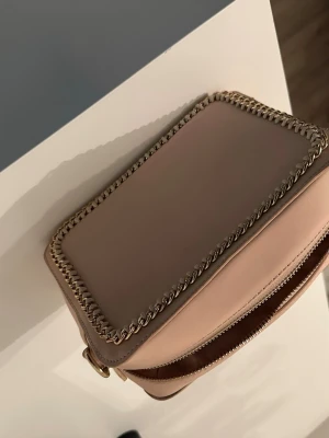 Beige axelväska med guldkedja - Snygg beige axelväska i boxig modell med guldfärgad kedja runt kanterna och som axelrem. Väskan har två fack med dragkedja och är gjord i slätt konstläder. Perfekt för dig som gillar stilrena detaljer och trendig design.