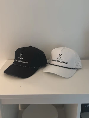 Svart och vit keps More Mulligans - Två kepsar från More Mulligans, en svart och en vit, båda med broderad logga och text framtill. Kepsarna har böjd skärm, ventilerande mesh på sidorna och justerbar baksida. Perfekt accessoar för en sportig och stilren look.
