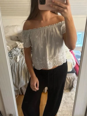 Vit offshoulder blus med spetsdetaljer - Söt vit offshoulder blus med tunna grå ränder och broderade blommor. Toppen har korta ärmar och avslutas med fina spetsdetaljer nertill. Perfekt för en fräsch och somrig look.