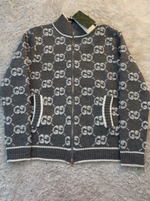 Gucci Cardigan - Snygg grå ziptröja från Gucci med vitt GG-mönster över hela tröjan. Tröjan har ribbade muddar, ståkrage och vita detaljer vid dragkedja, fickor och kanter. Perfekt för dig som gillar exklusiv streetwear och vill sticka ut.