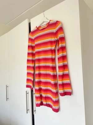Randig ribbad klänning i starka färger - Färgglad ribbad klänning med långa ärmar och rund hals. Mönstret är randigt i rosa, rött, orange, gult och vitt. Klänningen är kort och har en figurnära passform, perfekt för dig som gillar att sticka ut. Materialet känns mjukt och stretchigt.