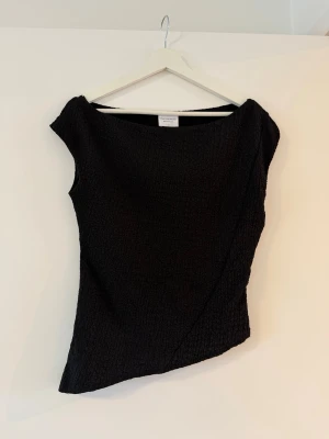 Svart topp  - Säljer en svart topp med asymmetrisk nederkant och strukturerat tyg. Toppen är ärmlös och har en bred halsringning som ger en cool och modern look. Perfekt att styla med jeans eller kjol för en trendig outfit.