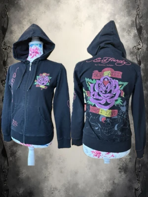 Svart Ed Hardy hoodie med tryck - Snygg svart hoodie från Ed Hardy med dragkedja och stor huva. Framsidan har ett färgglatt ros-tryck och logga, medan baksidan pryds av en stor lila ros, gröna blad och texten 'Dedicated One I Love'. Detaljer i rosa och grönt, samt ribbade muddar. Perfekt för dig som gillar streetwear och statement-plagg.