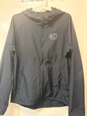 Svart Nike Air vindjacka med huva - Snygg svart vindjacka från Nike Air med dragkedja framtill och två sidofickor. Jackan har en huva och ett vitt Nike Air-logotyptryck på bröstet. Tillverkad i lätt och tunt material som passar perfekt för blåsiga dagar.
