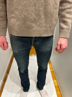 Mörkblå jeans från Nudie  - Säljer ett par mörkblå skinny jeans från Nudie Jeans med slitningar och hål på bakfickan. Jeansen har klassiska orange sömmar, fem fickor och är tillverkade i denim med stretch. Perfekta för en avslappnad och trendig look.