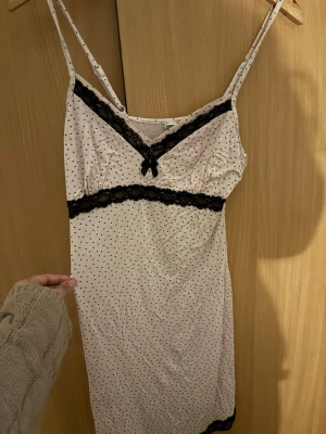 Ljusrosa nattlinne med spetsdetaljer - Sött ljusrosa nattlinne med tunna axelband och små svarta prickar. Dekorerat med svart spets vid bysten, midjan och nederkanten samt en liten rosett framtill. Mjukt och följsamt material som känns skönt mot huden.