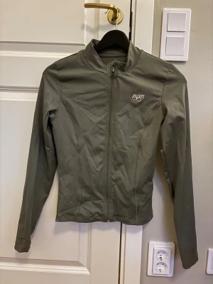 Olivgrön träningsjacka  - Snygg olivgrön träningsjacka från märket eight med dragkedja framtill och hög krage. Jackan har en slim passform och är tillverkad i ett lätt, syntetiskt material. 