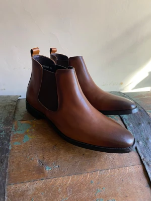 Bruna Chelsea boots i skinn - Snygga Chelsea boots i brunt skinn med klassisk siluett och elastiska sidopaneler. Bootsens ovandel är slät och har dragflikar bak för enkel påtagning. Perfekta för dig som vill ha en stilren och tidlös look.
