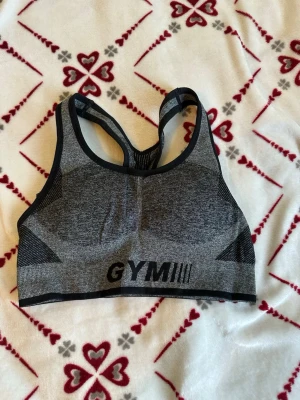 Grå sport-bh - Säljer en grå sport-bh  med svart kant och texten GYM framtill. Bh:n har brottarrygg och är tillverkad i ett stretchigt material som passar perfekt för träning. Materialet är mjukt och följsamt för extra komfort.
